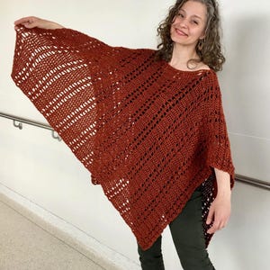 May include: A rust-colored, open-weave poncho. The poncho is made of a knitted material with a geometric pattern. The garment is draped over the shoulders and arms. The poncho is a warm, stylish accessory.