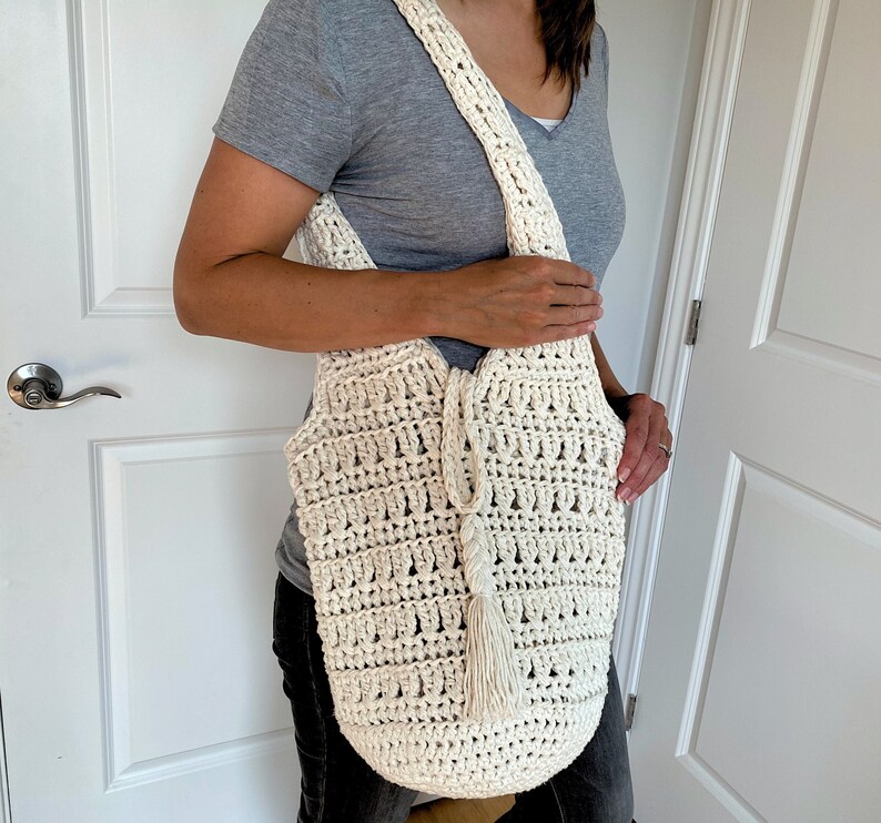 May include: A white crocheted tote bag with a long strap and a tassel. The bag is worn over the shoulder of a person wearing a gray shirt and jeans.