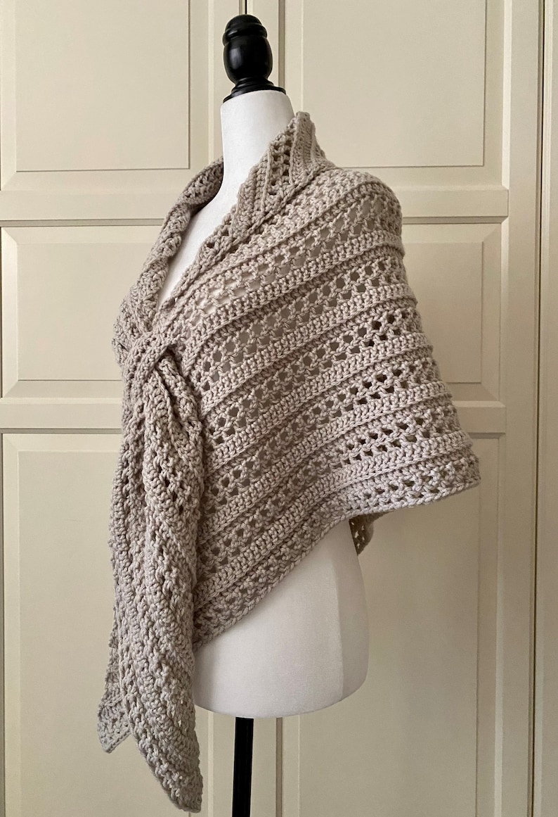 Peut inclure: Un ch&acirc;le en crochet beige avec un motif ajour&eacute; d&eacute;licat. Le ch&acirc;le est drap&eacute; sur un mannequin blanc et pr&eacute;sente un d&eacute;tail nou&eacute; au niveau du cou.