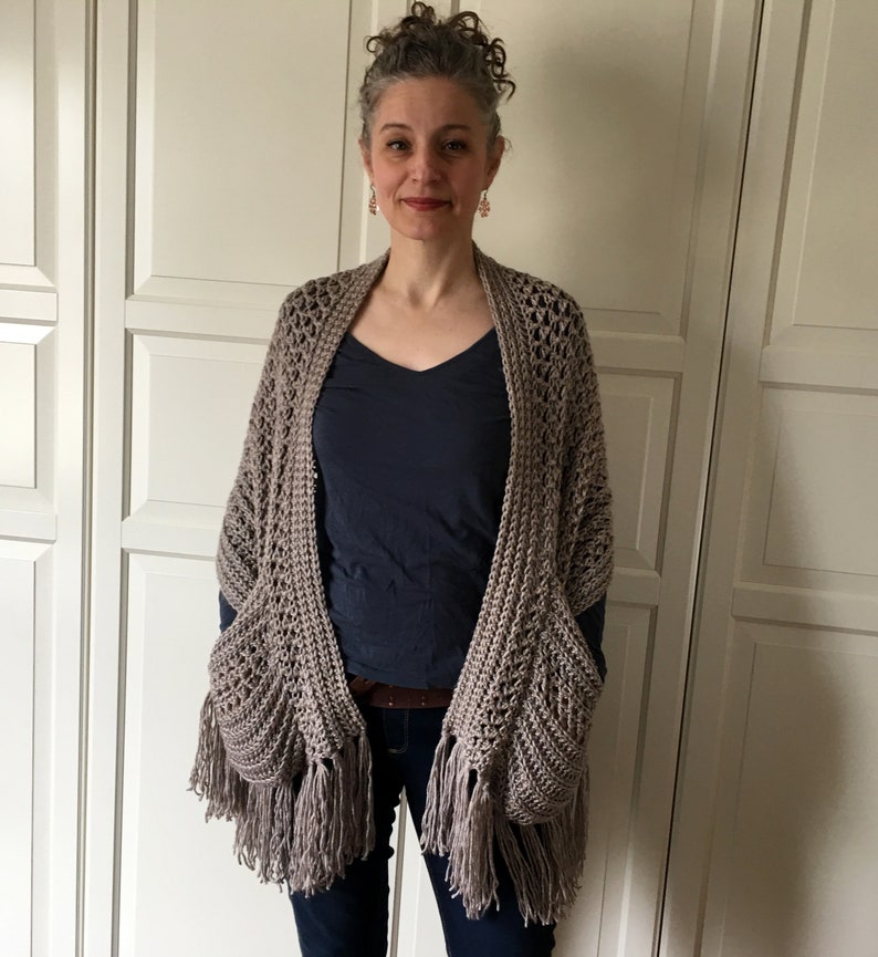 May include: A crocheted shawl with fringe and pockets, in a light brown color. The shawl is worn by a person with gray hair and a blue shirt.