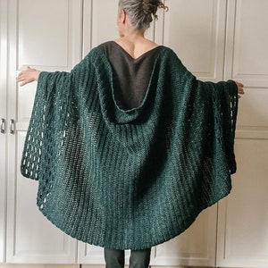 May include: A dark green crocheted hooded cape with a loose fit and a lacy pattern. The cape is worn by a person with their back to the camera.