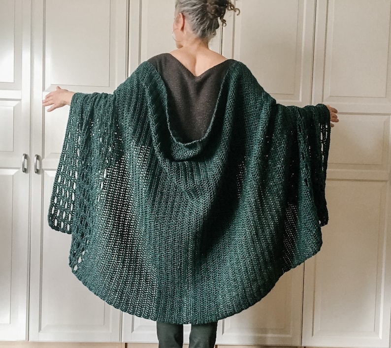 May include: A dark green crocheted hooded cape with a loose fit and a lacy pattern. The cape is worn by a person with their back to the camera.