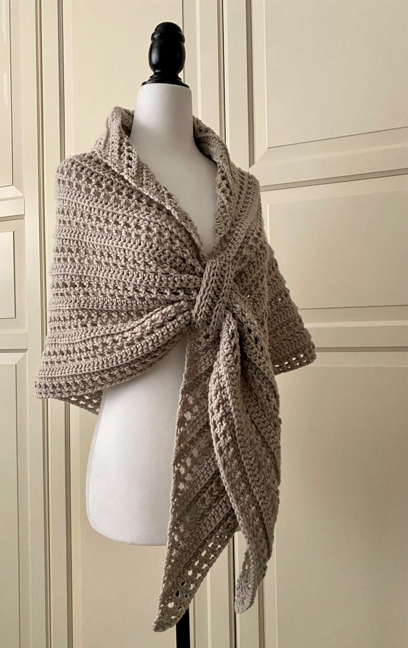 Peut inclure: Un ch&acirc;le en crochet beige avec un motif ajour&eacute; d&eacute;licat. Le ch&acirc;le est drap&eacute; sur un mannequin blanc et nou&eacute; au cou.