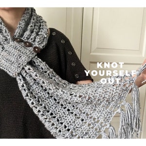 May include: A grey crocheted scarf with fringe and wooden buttons. The scarf is knotted around the neck and the text "KNOT YOURSELF OUT" is visible in the background.