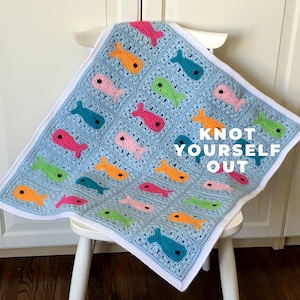 May include: A blue crocheted baby blanket with a white border featuring colorful fish designs. The blanket is draped over a white chair and the text "KNOT YOURSELF OUT" is visible in the lower right corner.