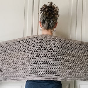 May include: A gray crochet shawl with fringe on the edges. The shawl is draped over the shoulders of a person.