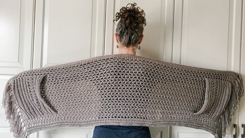 May include: A gray crochet shawl with fringe on the edges. The shawl is draped over the shoulders of a person.
