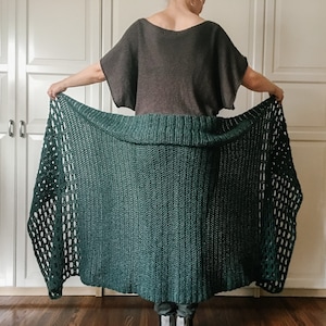 May include: A dark green crochet shawl with a lacy pattern. The shawl is draped over a person's shoulders and is being held in their hands.