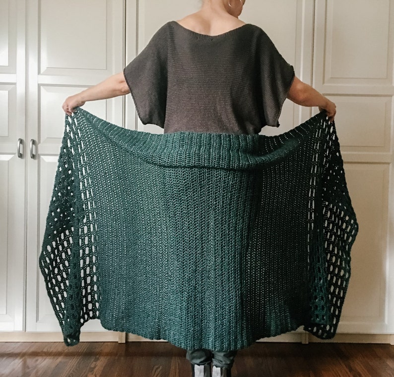 May include: A dark green crochet shawl with a lacy pattern. The shawl is draped over a person's shoulders and is being held in their hands.