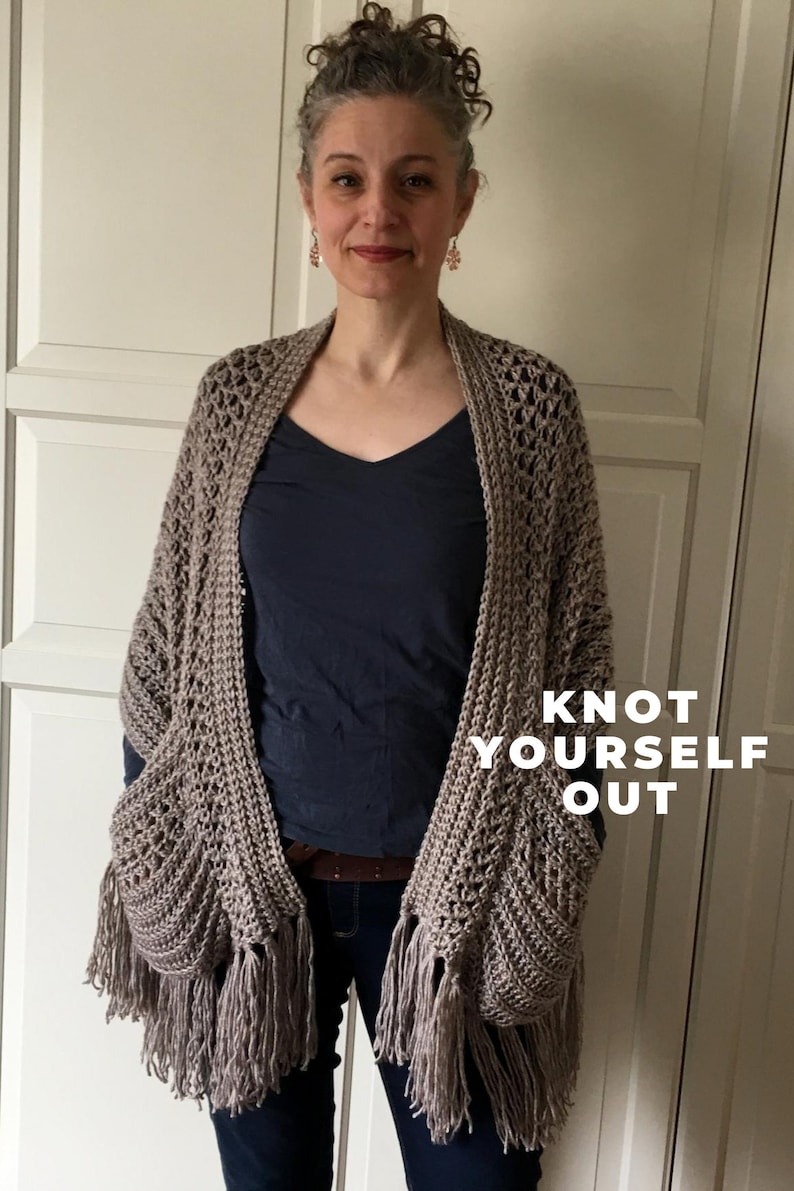 May include: A woman wearing a brown crocheted shawl with fringe. The shawl has two pockets and the text "Knot Yourself Out" is printed on the shawl.