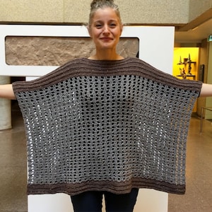 Puede incluir: Un poncho de ganchillo gris y marrón. El poncho tiene un diseño de tejido abierto y ancho con una banda marrón sólida alrededor del cuello y el dobladillo inferior. La persona lleva el poncho con los brazos extendidos.