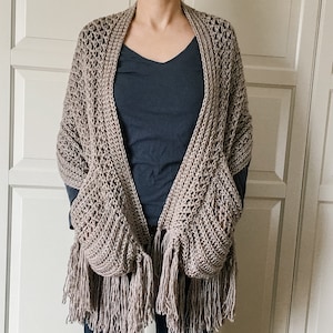 May include: A light brown crocheted shawl with fringe and pockets. The shawl is worn over a dark blue shirt and blue jeans.