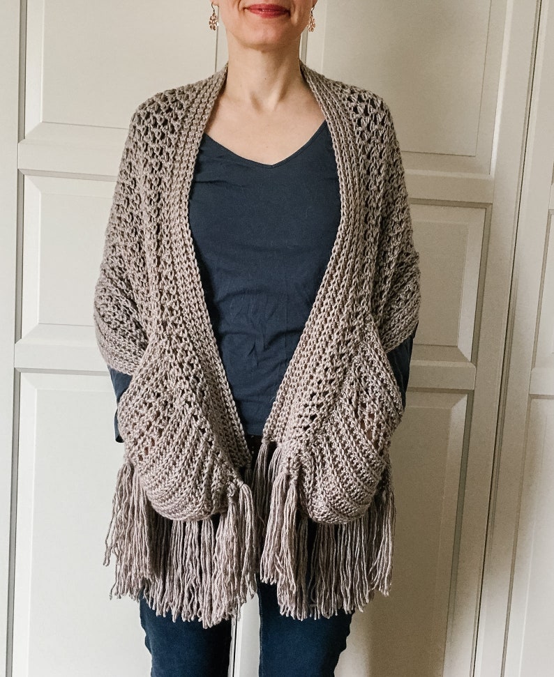 May include: A light brown crocheted shawl with fringe and pockets. The shawl is worn over a dark blue shirt and blue jeans.