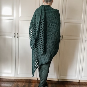 May include: A dark green crochet shawl with a lacy pattern. The shawl is draped over a person's shoulders and is worn with dark pants.