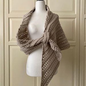 May include: A taupe-colored, crocheted shawl draped over a mannequin. The shawl features a textured stitch pattern and is tied at the front. The design includes openwork sections and a triangular shape, suggesting a cozy accessory.