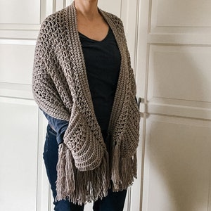May include: A brown crocheted shawl with fringe. The shawl is draped over a person's shoulders and has a loose, flowing fit.