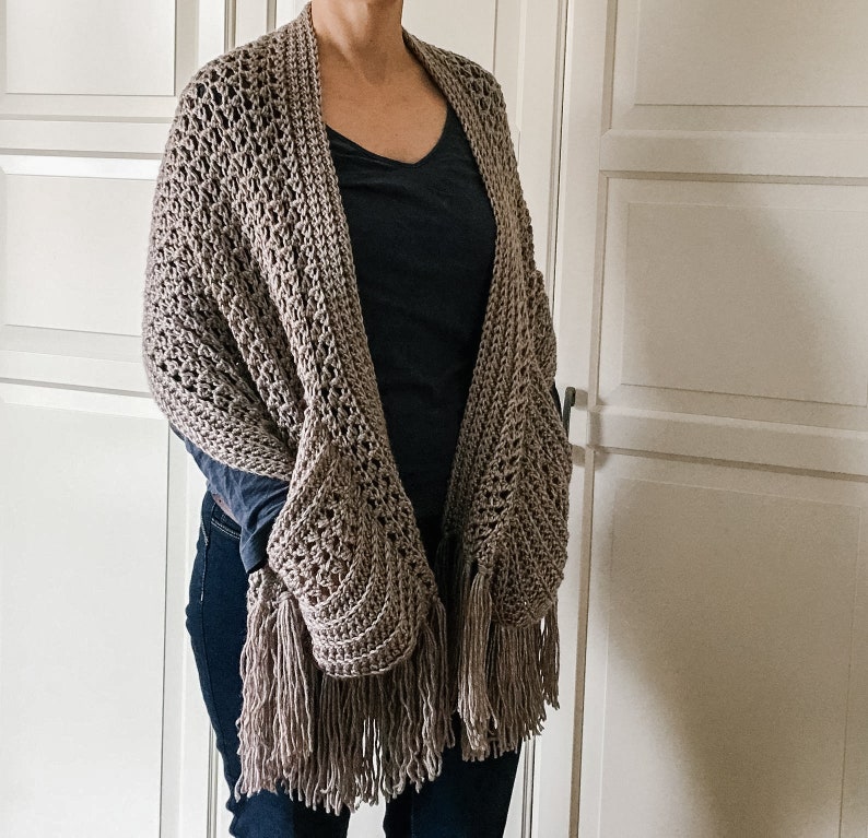May include: A brown crocheted shawl with fringe. The shawl is draped over a person's shoulders and has a loose, flowing fit.