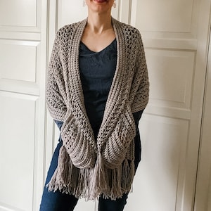 May include: A brown crocheted shawl with fringe, featuring a unique pocket design. The shawl is draped over a person's shoulders.