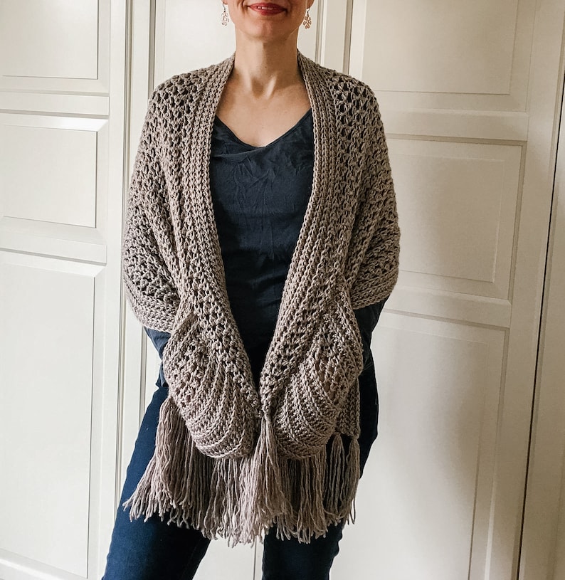 May include: A brown crocheted shawl with fringe, featuring a unique pocket design. The shawl is draped over a person's shoulders.