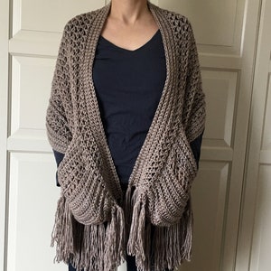 May include: A brown crocheted shawl with fringe and two pockets. The shawl is worn by a person with a dark blue shirt and dark blue jeans.