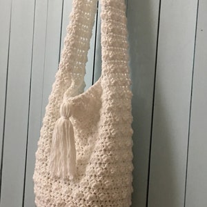 May include: White crocheted tote bag with a tassel hanging from the bottom. The bag is hanging on a light blue wall.