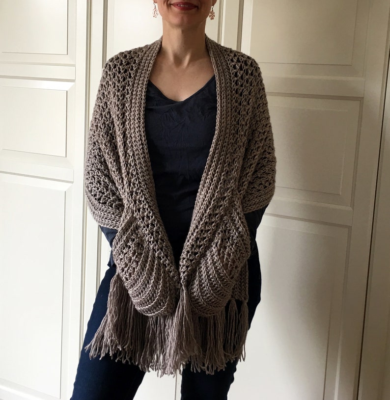 May include: A brown crocheted shawl with fringe. The shawl has a unique design with pockets on the sides.