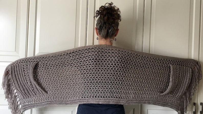 May include: A brown crocheted shawl with fringe on the edges. The shawl is draped over a person's shoulders.