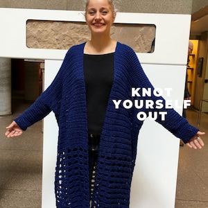 Könnte beinhalten: Eine Frau trägt einen langen, blauen Häkelcardigan mit einem Spitzenmuster am Saum. Der Cardigan ist vorne offen und die Frau trägt darunter ein schwarzes Shirt und eine blaue Jeans. Der Text "KNOT YOURSELF OUT" ist auf dem Cardigan sichtbar.