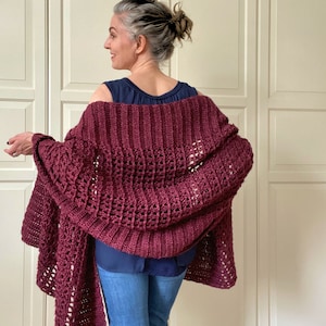 May include: A maroon crocheted shawl with a lacy pattern. The shawl is draped over the shoulders of a person wearing a navy blue tank top and blue jeans.