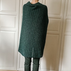 May include: A dark green knitted poncho with a cowl neck. The poncho is worn over dark green pants.