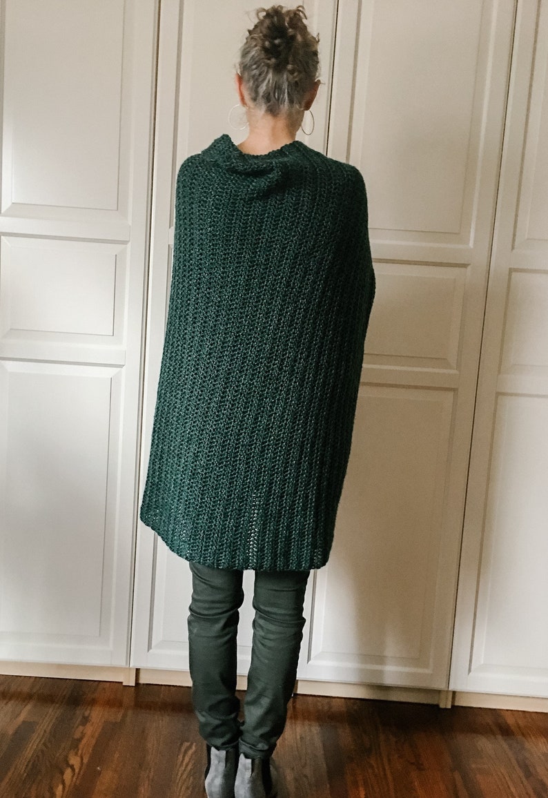 May include: A dark green knitted poncho with a cowl neck. The poncho is worn over dark green pants.