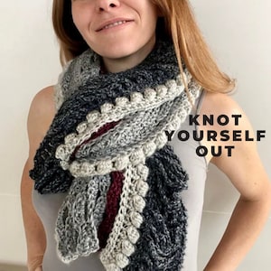 Puede incluir: Una mujer lleva una bufanda de ganchillo gris, blanca y burdeos con un diseño texturizado y pompones blancos. La bufanda está anudada alrededor de su cuello y el texto "Knot Yourself Out" es visible en la imagen.