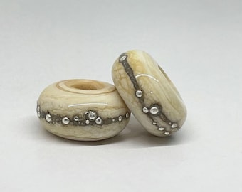 May include: Two round, cream-colored beads with a smooth, polished finish. Each bead features a textured, dark gray band encircling the center, embedded with small, raised, metallic silver spheres that create a distinct, tactile pattern.