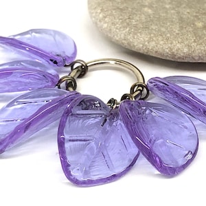 May include: A key ring with a silver-colored metal ring and seven translucent, purple, leaf-shaped charms. The charms are arranged around the ring, creating a floral or botanical theme. The background is white.