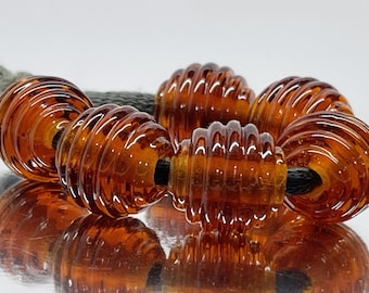 May include: A strand of translucent amber-colored glass beads, each featuring a distinct ribbed or beehive-like texture. The beads are strung together on a dark cord, creating a repeating pattern of rounded, grooved shapes with a glossy finish.