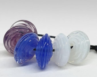 May include: A collection of five circular glass beads strung on a black cord. The beads feature a ribbed, spiral texture and include translucent purple, vibrant blue, and opaque white colors, each with a central hole for threading.
