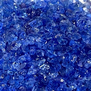 May include: Close-up of a pile of small, irregularly shaped, translucent blue glass pieces. The pieces are reflective, catching the light and creating a sparkling effect. The overall color is a deep, vibrant blue.