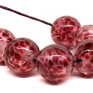 May include: Close-up of a strand of round, glass beads in shades of burgundy and pink. The beads have a textured, speckled appearance with swirling patterns. The beads are strung on a dark brown cord, perfect for jewelry making.
