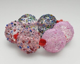 May include: A bracelet composed of six spherical glass beads with textured surfaces. The beads feature a mix of pink, blue, green, and clear glass fragments, connected by a bright red cord that is visible between each individual bead.
