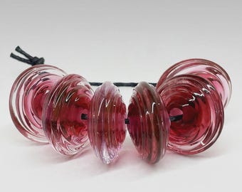 May include: A collection of five translucent, deep magenta glass beads with a spiraled, ribbed texture. The circular beads are strung together on a thin black cord, showcasing a glossy finish and a layered, circular design throughout each piece.