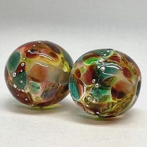 May include: Two spherical glass beads featuring a multicolored pattern of swirling amber, teal, and green tones. The surface is decorated with small, raised silver-colored metallic dots embedded within the clear glass structure.