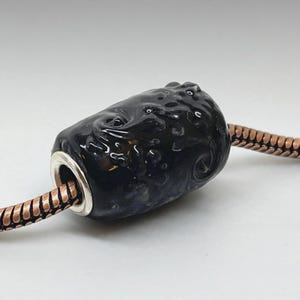 May include: A cylindrical black glass bead with a glossy, textured surface featuring swirling patterns and raised bumps. The bead has a silver-toned metal core and is threaded onto a copper-colored snake chain necklace with a metallic finish.