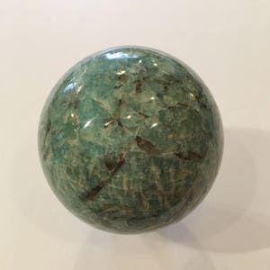 May include: A green and white sphere made of polished amazonite. The sphere has a smooth, polished surface with a natural, marbled pattern.