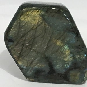 May include: A large, dark green, iridescent labradorite gemstone with a hexagonal shape. The stone has a smooth, polished surface and displays a variety of colors, including blue, green, and gold.