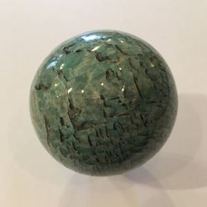 May include: A green, polished, spherical stone with a marbled pattern. The stone is smooth and has a natural, organic look.