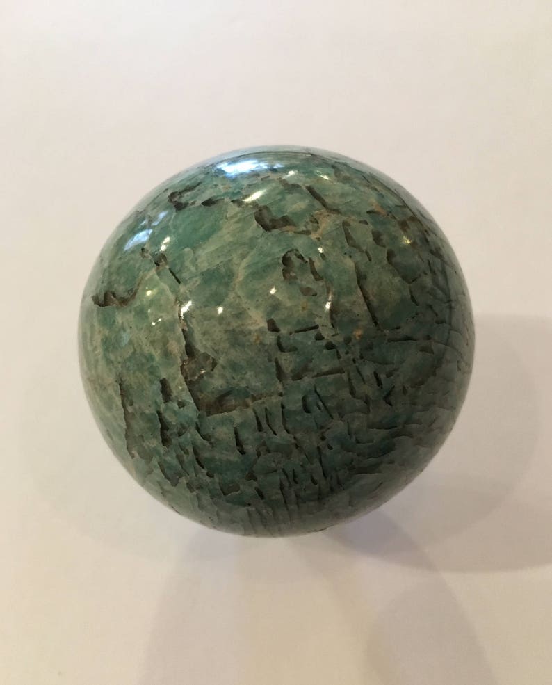 May include: A green, polished, spherical stone with a marbled pattern. The stone is smooth and has a natural, organic look.