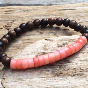 Puede incluir: Un brazalete hecho de cuentas de madera marrón oscuro y cuentas de coral rosa claro.