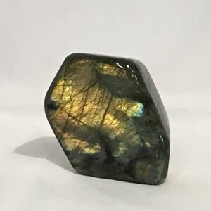 May include: A polished, dark green and gold labradorite gemstone with iridescent flashes. The stone is shaped like a hexagon and has a smooth, polished surface.