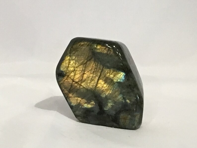 May include: A polished, dark green and gold labradorite gemstone with iridescent flashes. The stone is shaped like a hexagon and has a smooth, polished surface.