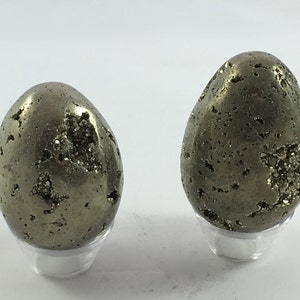 May include: Two pyrite eggs, one larger than the other, are displayed on clear stands. The eggs are a metallic gold color and have a rough, textured surface.